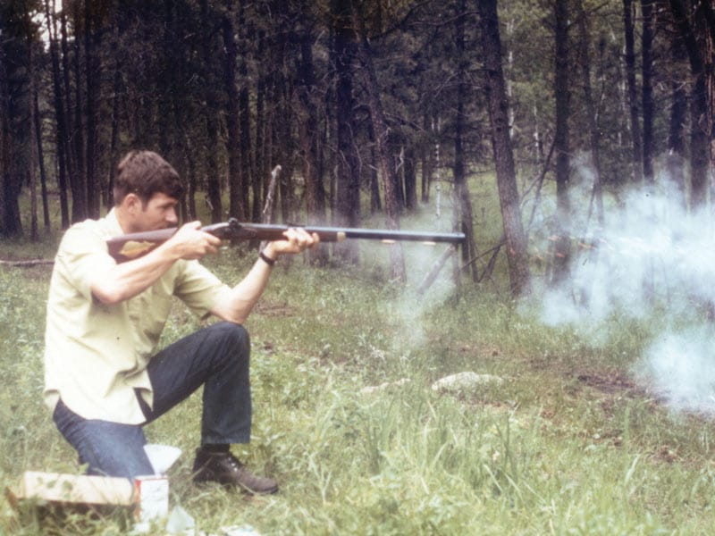Larry crouching and firing a Thompson/Center Hawken in the Black Hills of South Dakota with a white puff of smoke to the right.